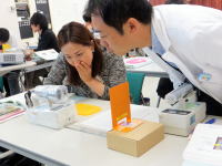 医療の研修中、男性講師が女性受講者に機器の操作を説明している様子。テーブル上には医療機器や資料が並んでいる。