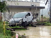 倒壊した建物の近くに停まる損傷した車両の様子。