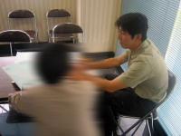 健康相談の場面で、テーブルを挟んで話し合う二人の男性が写っている。背景には椅子と窓が見える。