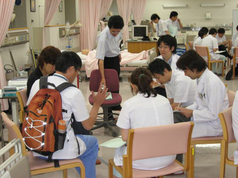 臨床実習中の学生たちがグループでディスカッションを行っている様子。背景にはベッドや医療機器が見える。