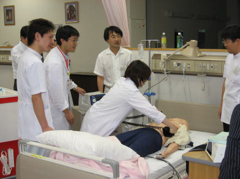 医学生たちがクリニカル・シナリオの実演を行っている様子。ベッドには人形が置かれ、学生が協力して医療行為を学んでいる。