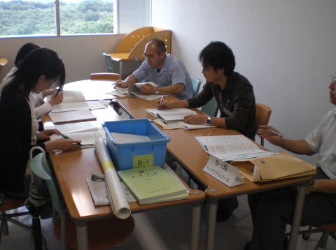 テーブルを囲んで資料を見ながら話し合う5人の参加者がいる会議の様子。
