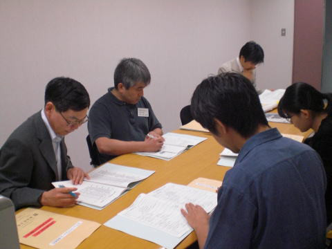 テーブルに向かって資料を読みながら作業する数人の人々の様子。