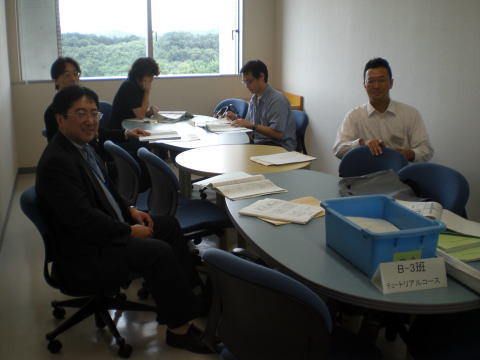 会議室で資料を見ながら話し合う人々の様子。テーブルには書類や文房具が散らばっている。
