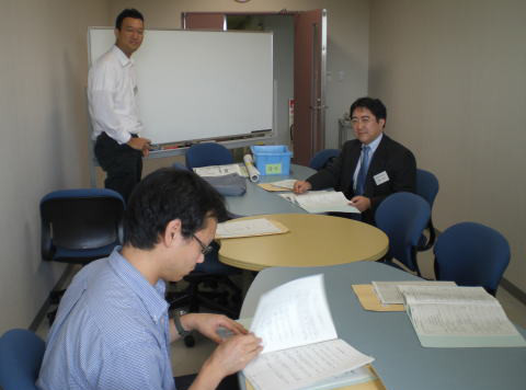 会議室で資料を確認している3人の男性が写っている様子。テーブルには書類が広げられ、ホワイトボードも見える。