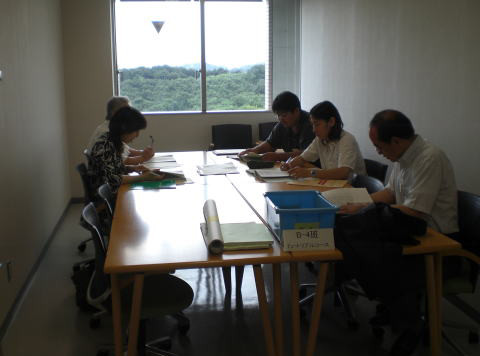 会議室で資料を見ながら話し合う5人の人々。窓からは緑の景色が見える。
