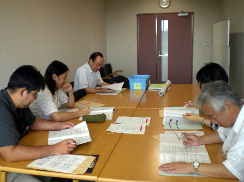 テーブルを囲んで資料を読み込む5人の参加者がいる会議室の様子。