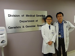 医療遺伝学部門の看板の前で白衣を着た二人の医師が並んで立っている様子。