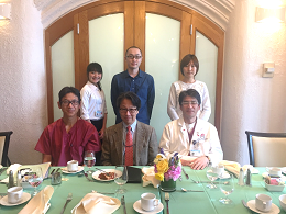 海外研修のために集まった医療関係者のグループ写真。テーブルには食事と花が飾られ、背景には木製のドアが見える。