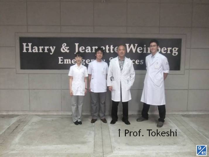 医療現場で白衣を着た4人の医師が、病院の看板の前でポーズをとっている様子。