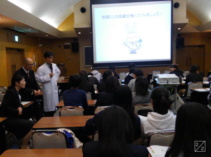 学校見学の様子。医療に関する説明を受ける学生たちと、講演者が映る教室の風景。