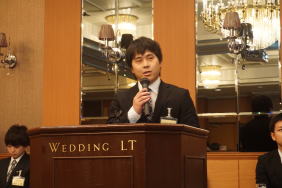 福島県立医科大学附属病院の臨床研修医が講演を行っている様子。背景にはシャンデリアが輝く会場。