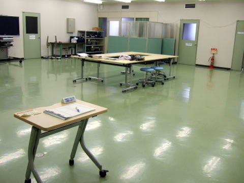 オープンスペースの教室内、机や椅子が配置され、明るい照明が当たる静かな環境。