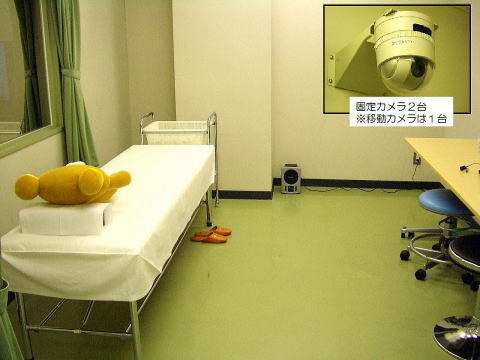 診察室の内部、ベッドの上にクマのぬいぐるみが置かれ、壁には監視カメラが設置されている様子。