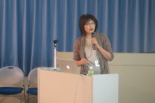 心エコーセミナーで講演を行う女性が、プレゼンテーションを行っている様子。背景には青いカーテンがあり、机の上にはパソコンと飲み物が置かれている。