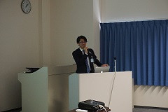 腹部エコーセミナーで講演を行う男性が、プレゼンテーションを行っている様子。背景には青いカーテンと時計が見える。