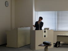 腹部エコーセミナーで講演を行う女性が、プレゼンテーションをしている様子。背景には時計と窓が見える。