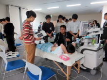 腹部エコーセミナーでの実習風景。受講者が患者にエコー検査を行い、周囲には見学者が集まっている。