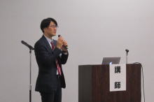 腹部エコーセミナーで講演する講師がマイクを持ち、聴衆に向けて話している様子。
