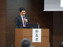 発表者が講演を行っている様子。背景には「発表者」と書かれた掲示があり、会場の参加者が注目している。