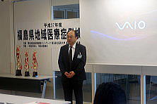 福島県地域医療症例検討会で講演する男性が立っている様子。背景には「福島県地域医療症例検討会」の看板とトロフィーが見える。