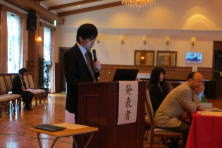 福島県地域医療症例検討会で発表する研修医の磯松大介先生。壇上でマイクを持ち、聴衆に向けて話している様子。