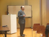 心電図セミナーで講演する男性が、聴衆に向けて話している様子。背景にはホワイトボードと椅子が並んでいる。
