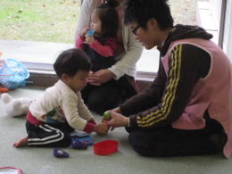 子どもと遊ぶ男性が、玩具を使って交流している様子。背景には他の子どもと大人が見える。