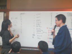 参加者がホワイトボードに向かい、意見を共有している様子。講師がマイクを持ち、説明を行っている。