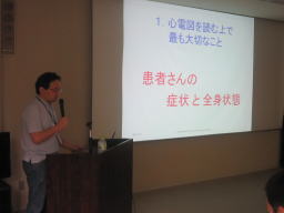 心電図に関する講義を行う講師と、スクリーンに表示された「患者さんの症状と全身状態」のスライド。