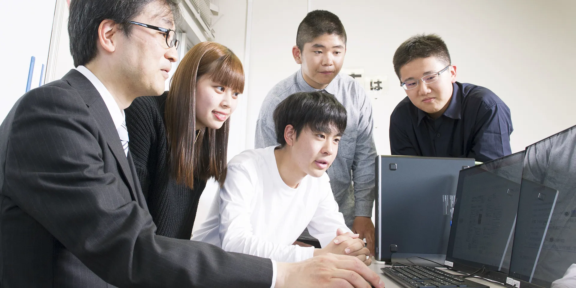 大学の教員と学生がパソコンを囲んで話し合い、研究や学習に取り組んでいる様子。