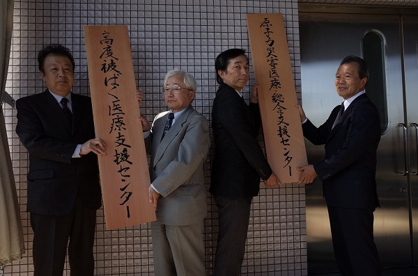 原子力災害医療・総合支援センターの開所式で看板を持つ4人の関係者。