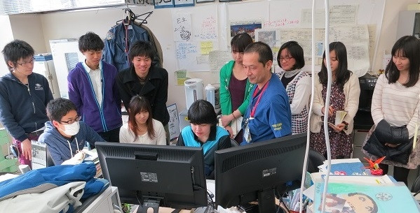医学生たちがコンピュータの前で研修を受けている様子。周囲には指導者や他の学生が集まっている。