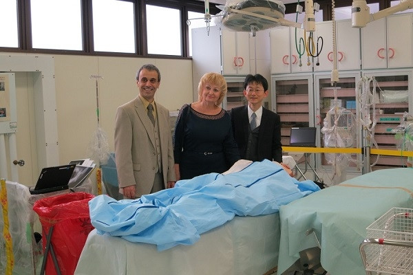 医療施設内で、3人の人物が手術台の前で笑顔でポーズをとっている様子。