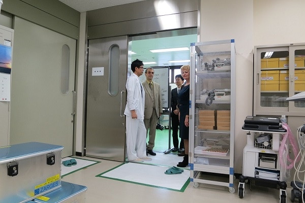 医療施設の入口で、白衣を着た医師と訪問者が話し合っている様子。背景には医療機器や収納棚が見える。