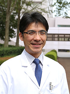 福島県立医科大学の丸橋繁教授が白衣を着て微笑んでいる写真。背景には大学の建物と緑の木々が見える。