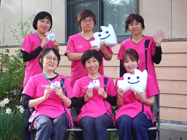 訪問看護ステーションすかわのスタッフが笑顔で記念撮影をしている様子。ピンクのユニフォームを着用し、手に白いマスコットを持っています。