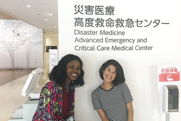 災害医療高度救命救急センターの前で笑顔の女性2名がポーズをとっている写真。