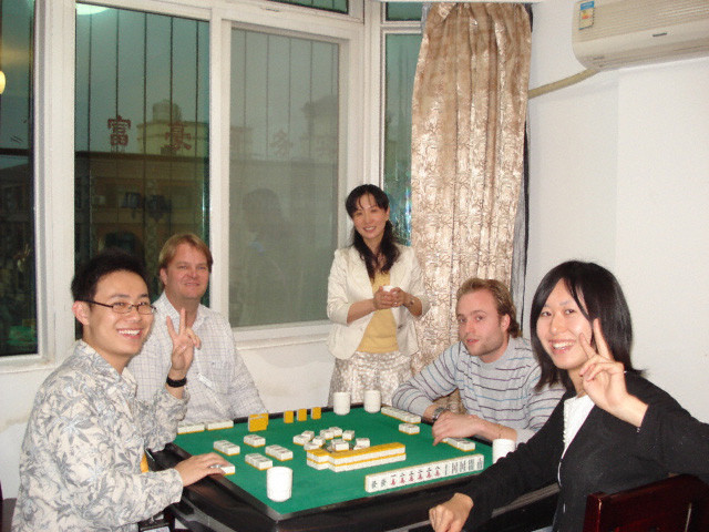 室内で5人の人々が麻雀を楽しんでおり、うち2人がピースサインをしています。リラックスした雰囲気の中で社交的な時間を過ごしています。