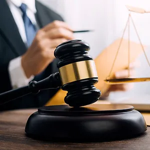 法廷のハンマーと天秤が置かれた机の上で、スーツを着た人物が書類に目を通している様子。