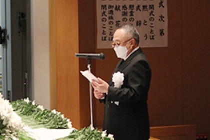 理事長がマイクの前でスピーチをしている様子。背景には祝辞の書かれた掲示物が見える。