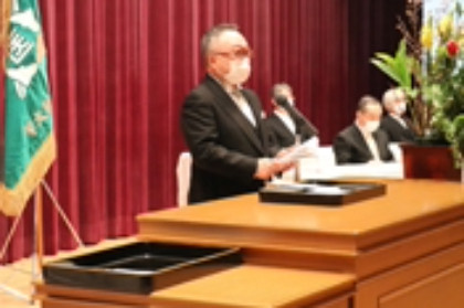 福島県立医科大学の理事長が壇上でスピーチを行っている様子。背景にはカーテンと装飾が見える。