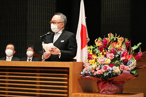 理事長がマスクを着用し、花束の前でスピーチを行っている様子。背景には日本の国旗が見える。