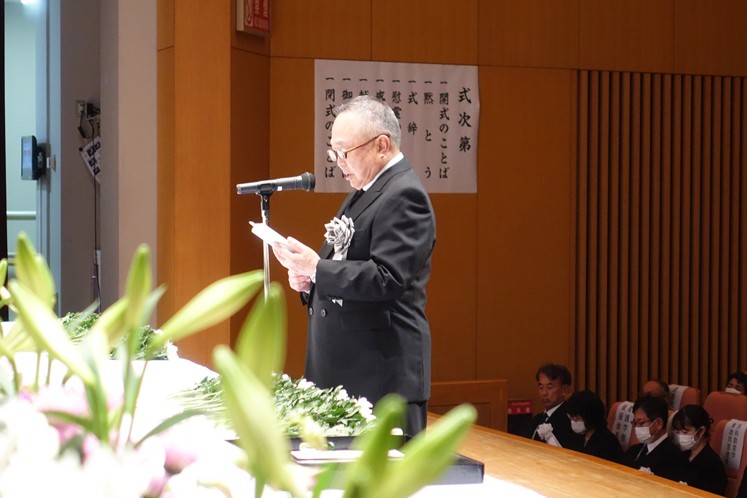 解剖慰霊祭でスピーチをする男性が、壇上で資料を手に持ちながら話している様子。背景には花や参加者の姿が見える。