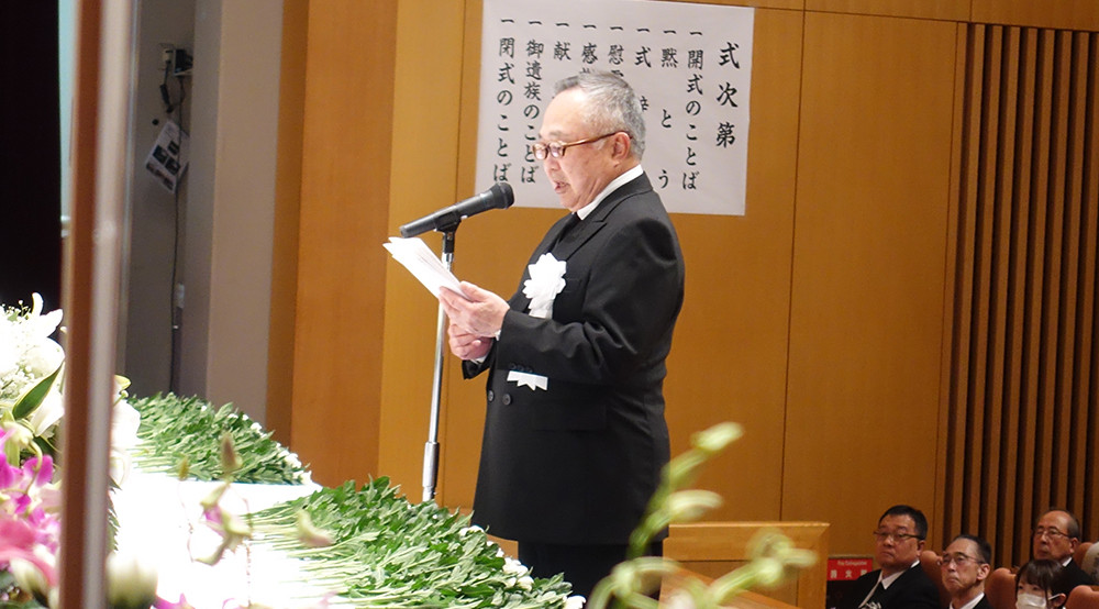 解剖慰霊祭でスピーチをする男性が、壇上で資料を手に持ちながら話している様子。背景には花や参加者の姿が見える。