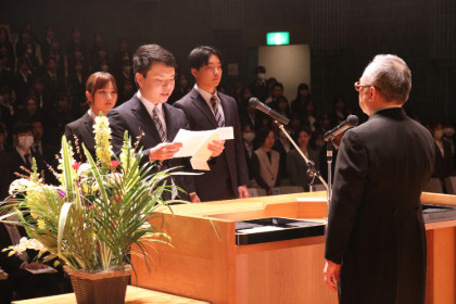 入学式でスピーチをする学生とそれを見守る教員、背景には多くの参加者がいる様子。
