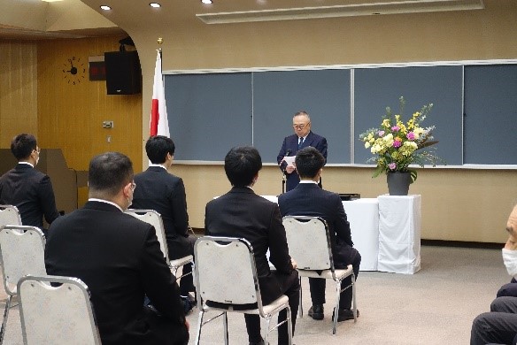 大学院医学研究科の入学式で、壇上で挨拶する教授と、前方に座る新入生たちの様子。背景には日本の国旗と花が飾られている。