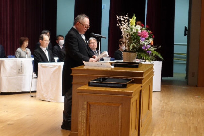 学位記授与式でスピーチをする男性と、花が飾られた壇上の様子。