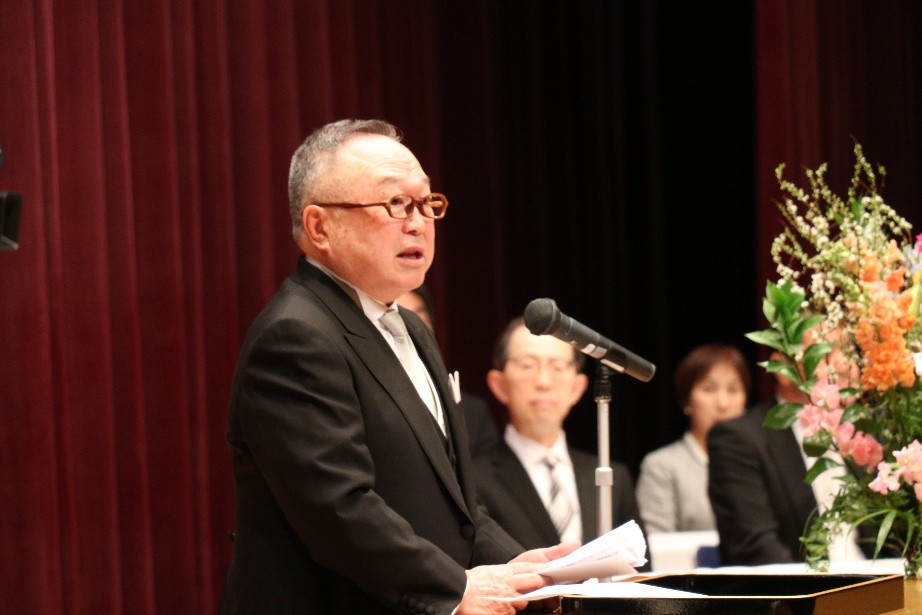 学位記授与式でスピーチをする理事長と、花が飾られた壇上の様子。