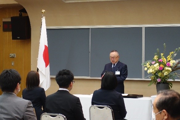 大学院の学位記授与式でスピーチをする男性と、参加者たちが座っている様子。背景には日本の国旗と花が飾られている。